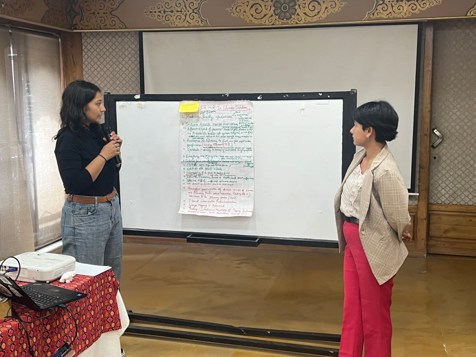 Two women stand in front of a whiteboard during a workshop presentation. One holds a microphone while the other listens attentively. A handwritten poster with discussion points is displayed between them.