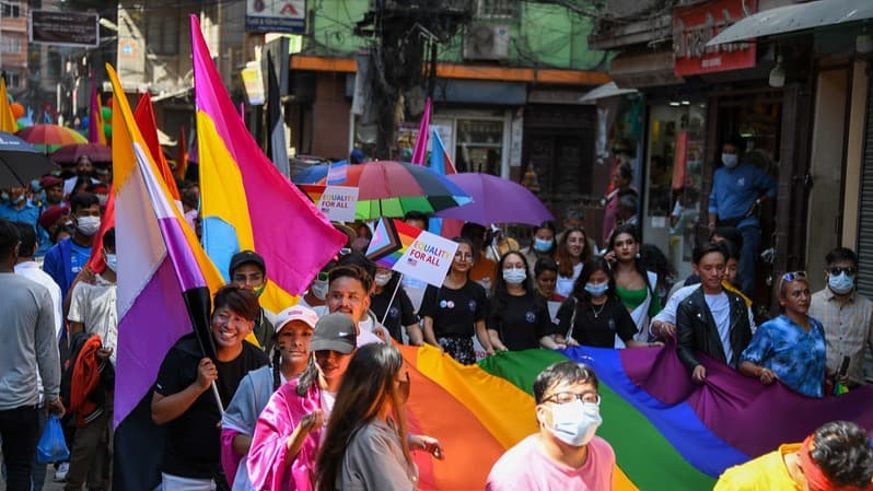 A crowd of diverse individuals, some holding various LGBTQ+ flags and rainbow umbrellas, parade down a narrow street with buildings on either side.