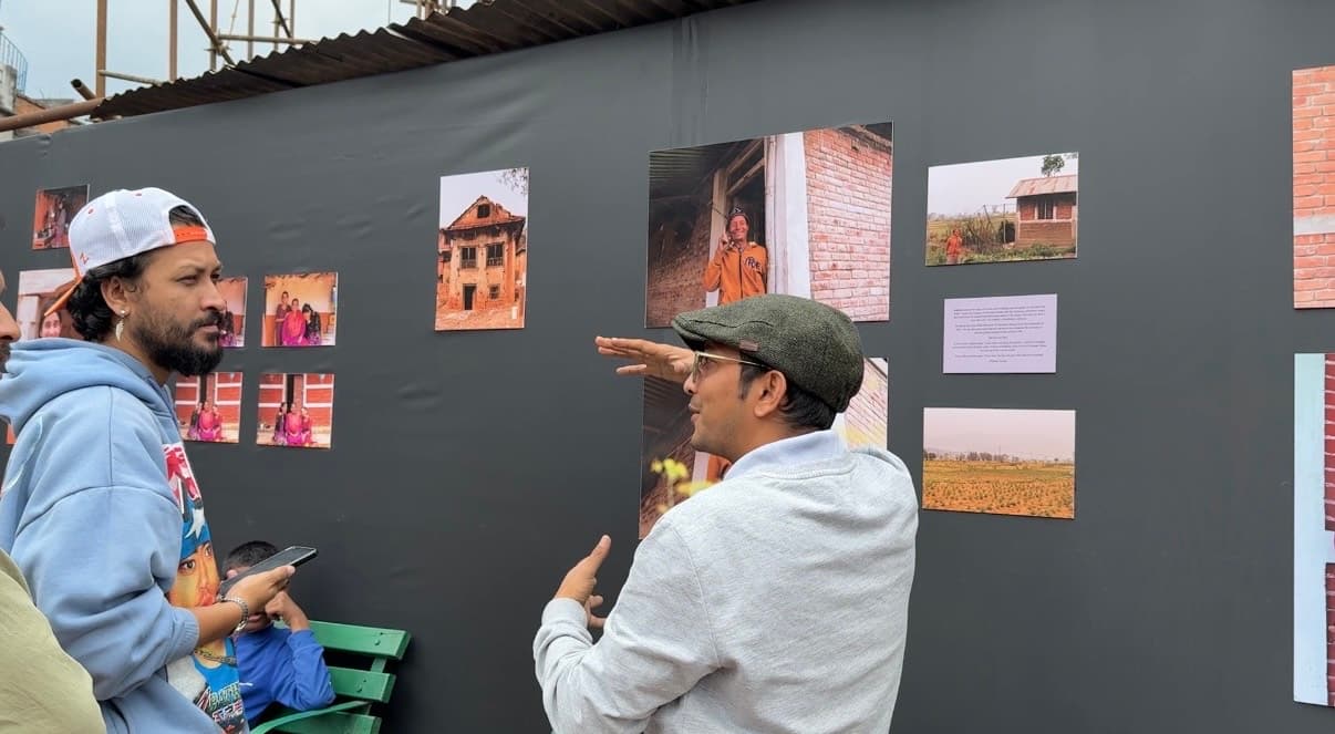 Three people are looking at photographs displayed on a dark gray wall, with one person gesturing towards a portrait of a smiling individual.