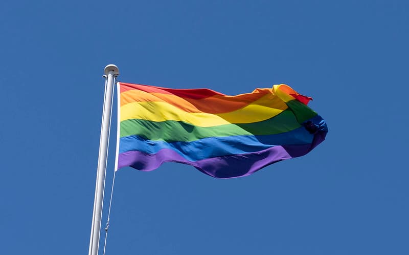 A vibrant rainbow pride flag waves against a clear blue sky, attached to a white flagpole on the left side of the frame.