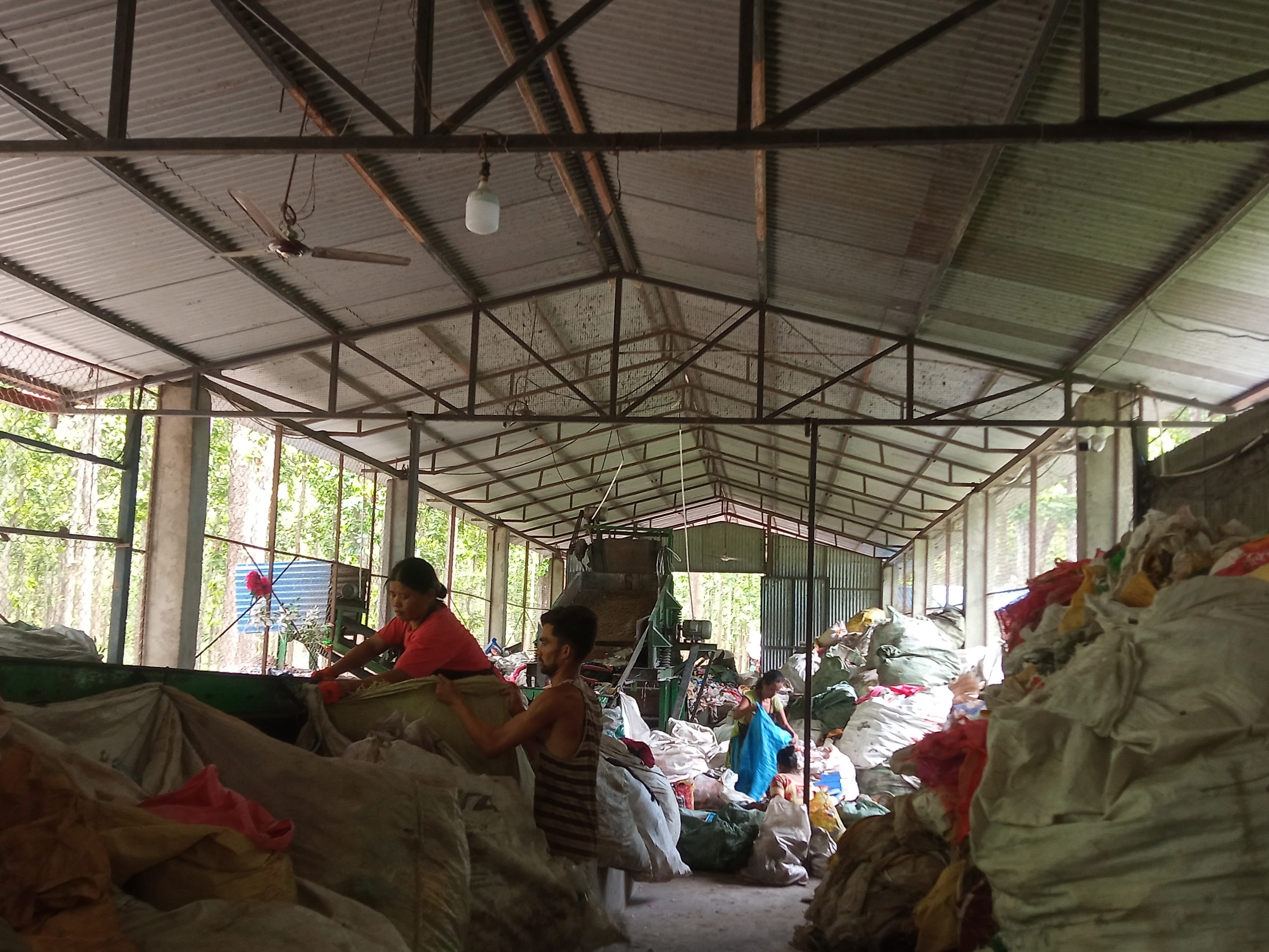 Butwal MRF's interior shows workers actively sorting plastic waste at a conveyor, with large sacks stacked around. One man and woman are seen carrying a sack. Ceiling fan and light are on the left.