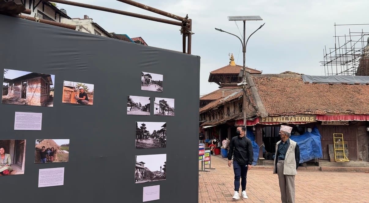 A photo exhibit on a dark gray wall stands in a public square with traditional brick buildings and people walking in the background.