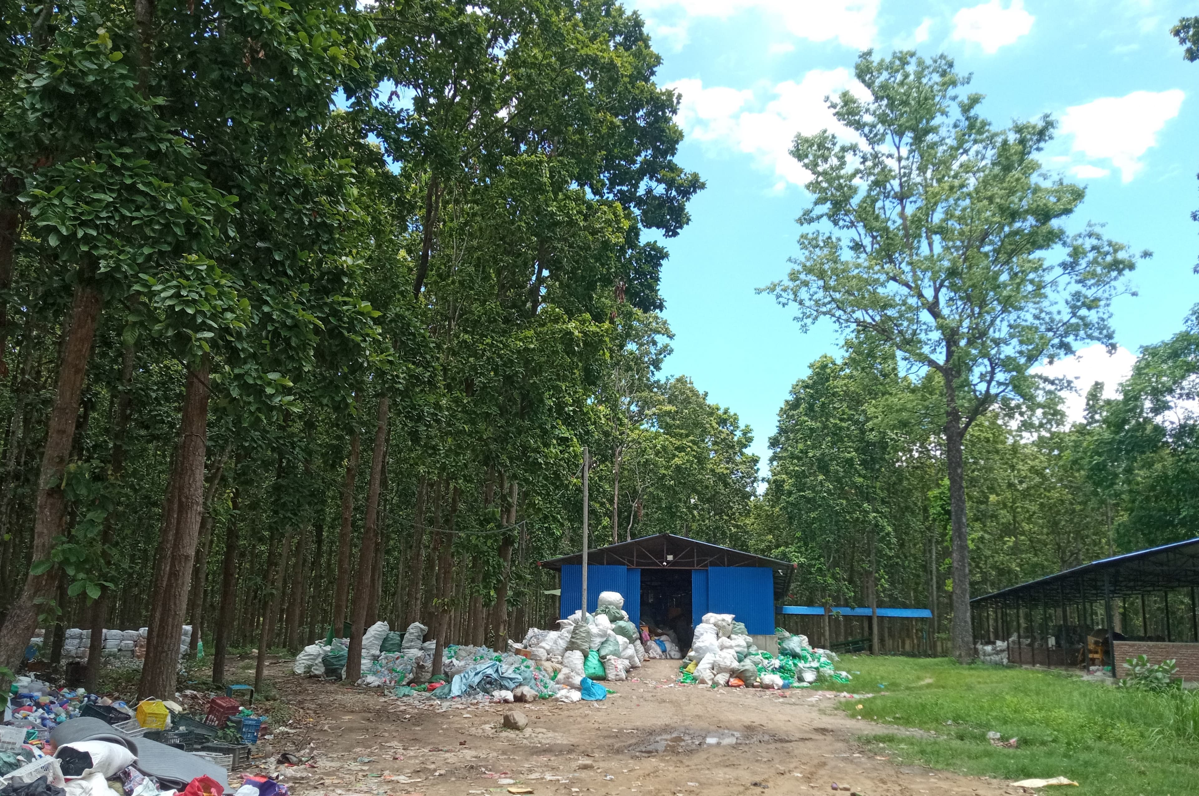 A blue truss Material Recovery Facility (MRF) sits within a Sal forest, flanked by two sheds. White sacks of segregated plastic bottles are visible at the entrance and inside, with some plastic waste scattered nearby.